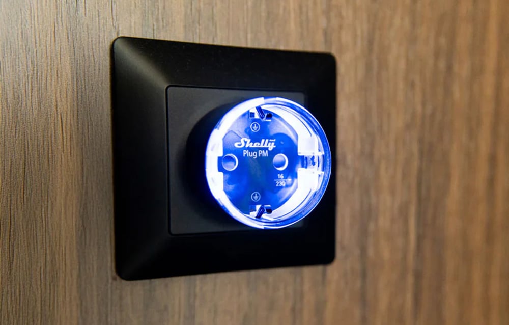 Shelly Plug PM smart socket with blue LED backlight mounted in a black frame on a wooden wall.