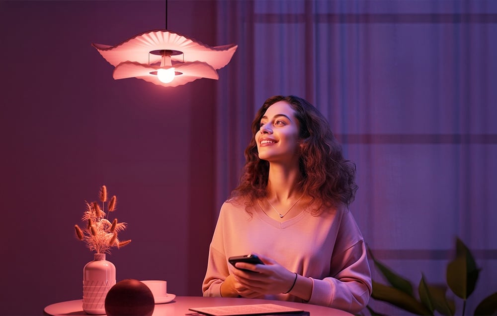 A smiling woman sitting at a table holds a phone, looking up at a glowing pink light bulb.