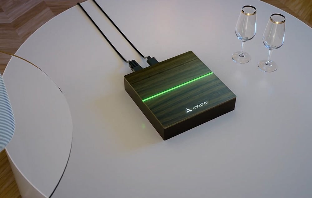 The square Matter device with wooden casing and green illumination in the middle stands on a white round table next to two glasses