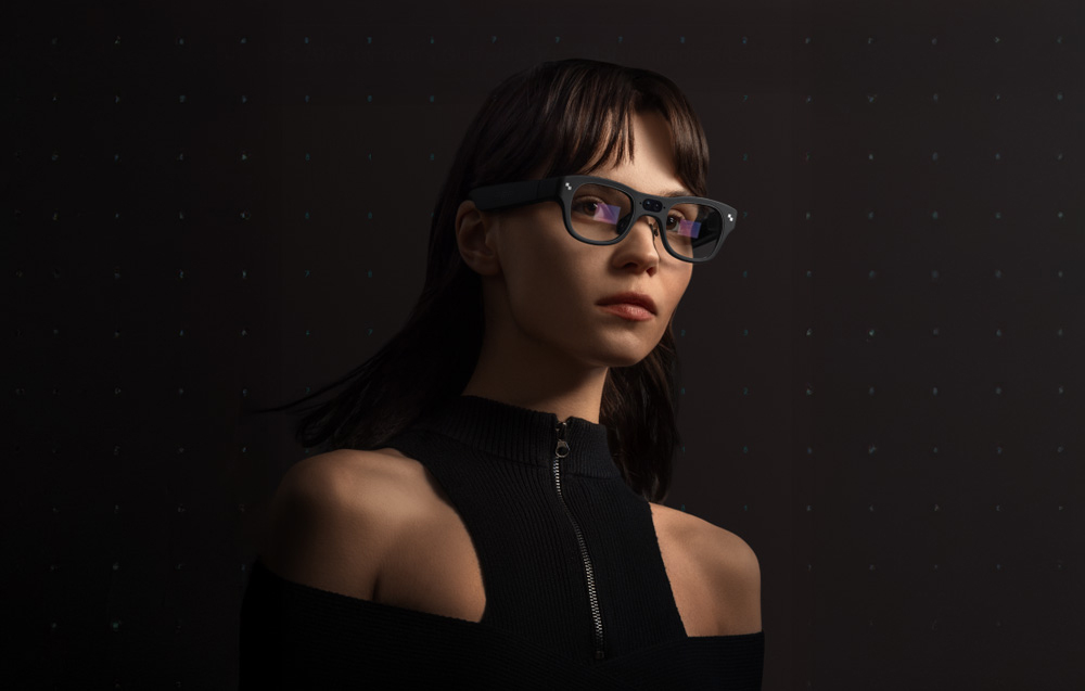 Woman in dark clothing wearing AR glasses against a dark wall with a subtle pattern.