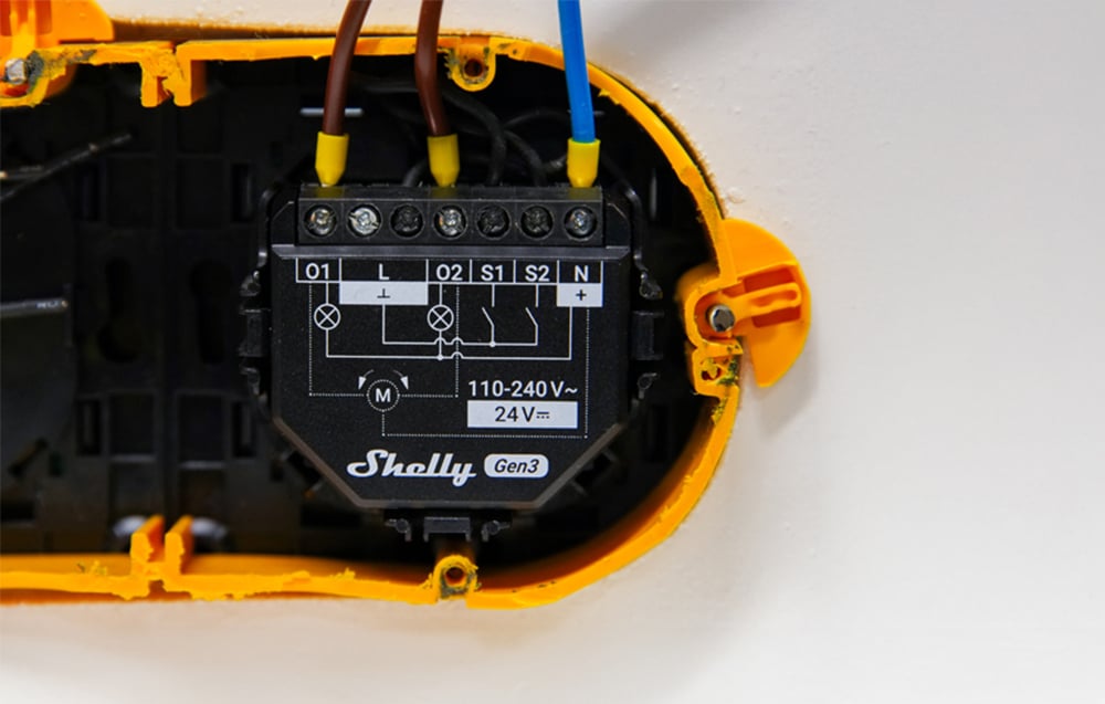Shelly 2PM Gen3 module mounted in electrical box with wires connected, surrounded by yellow mounting hardware