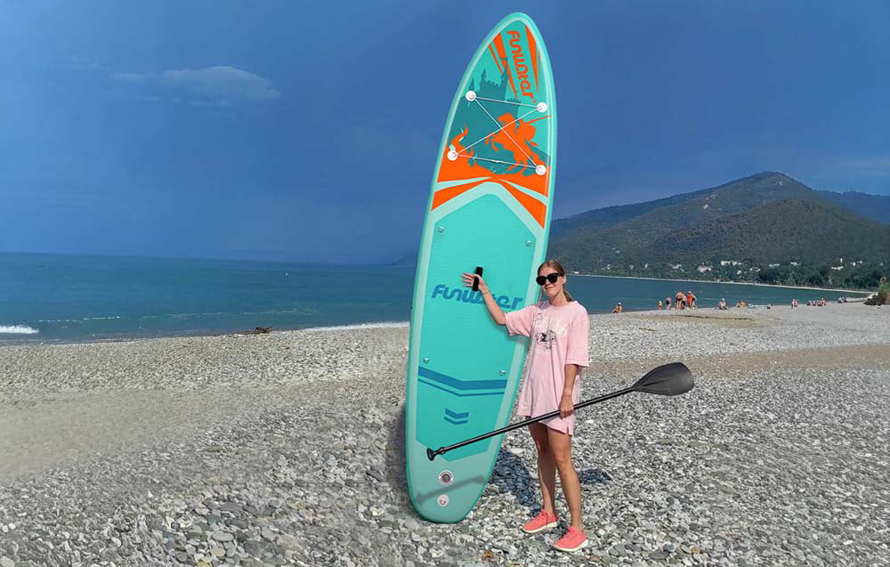 A young woman wearing sunglasses stands on a rocky beach holding a surfboard and paddle in her hands