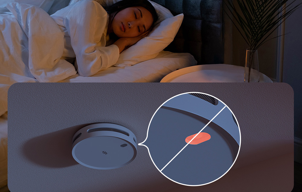 A smoke detector on the ceiling with the visual alarm turned off; a woman is sleeping in the room