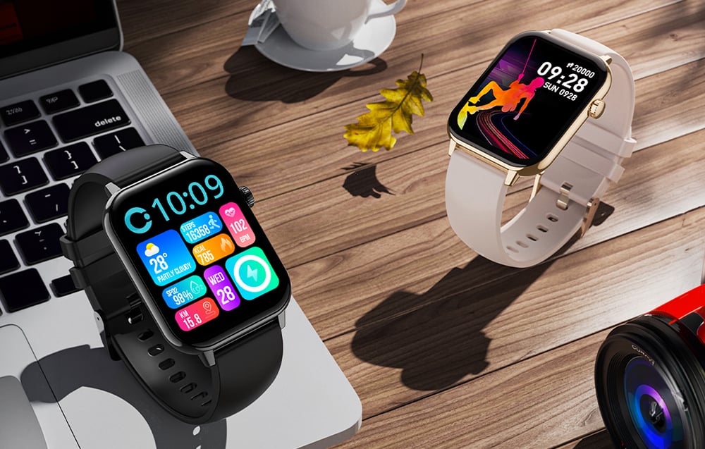 Two Colmi smartwatches (black and gold) on a wooden desk with a laptop and coffee