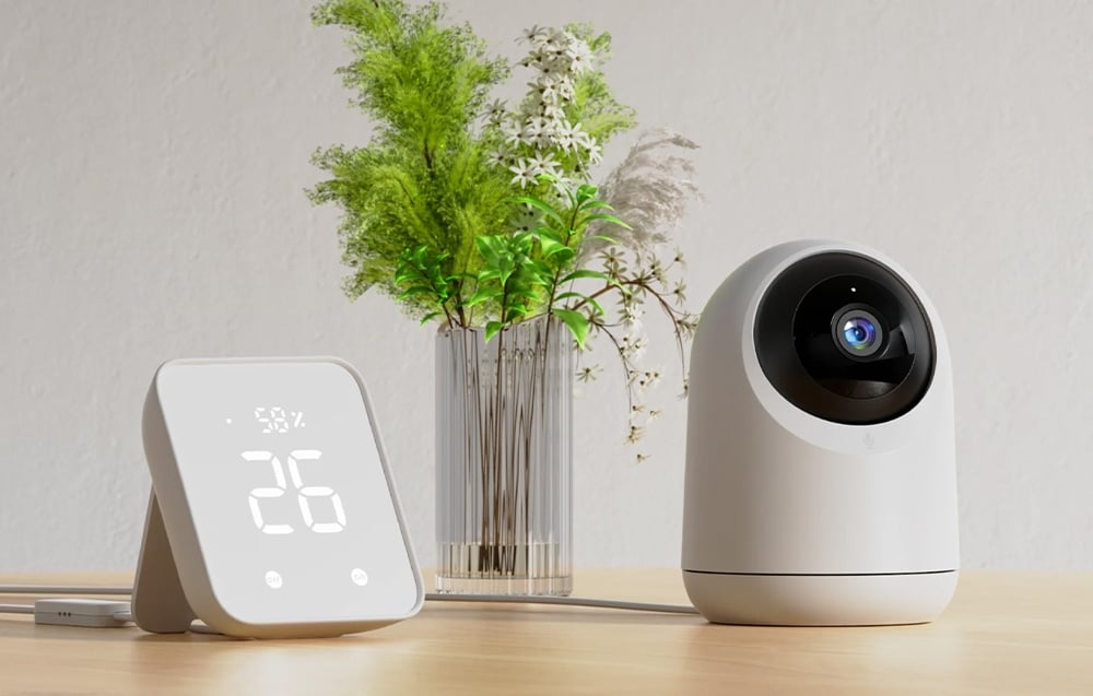 A white pan-tilt camera and an electronic room thermometer standing next to it on a table beside a vase with green plants.